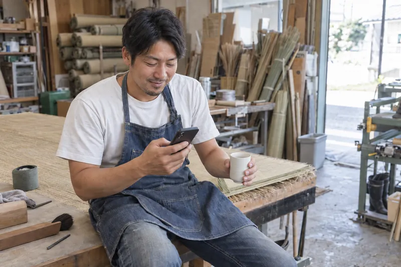 作業台に腰かけ湯のみを手にスマホを見る職人